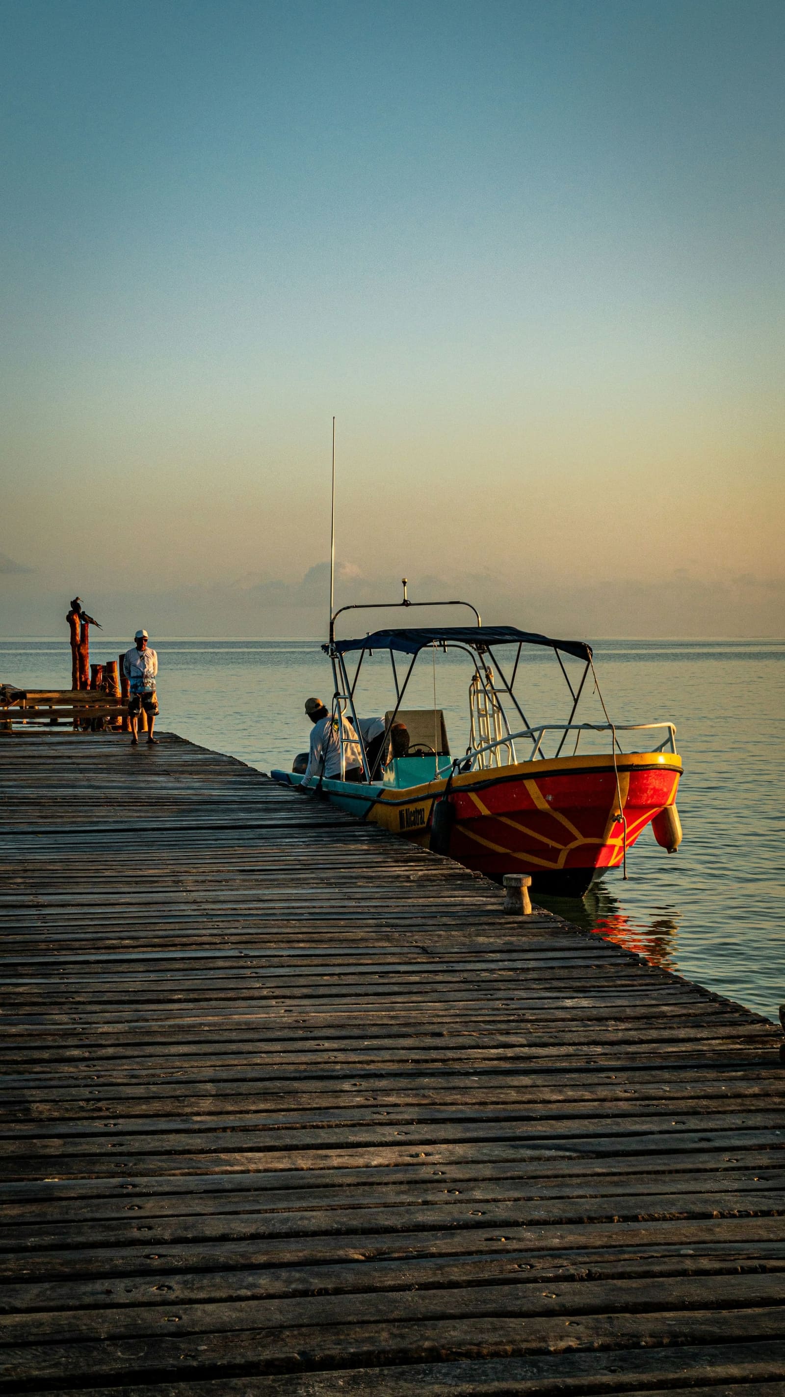 A wooden dock running out to a skiff tied up on flat water in the late afternoon